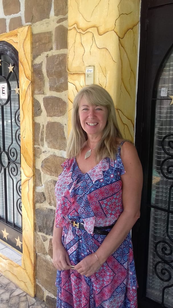 Hi, I'm Dee Lynn. I retired at 50. In this photo, I am standing in front of a yellow wall. The photo is of me from head to thigh and I am wearing a blue/pink/purple summer dress. It has a thin black belt. I also am wearing a necklace with a silver leaf pendant. My oval face is framed by medium length blonde grey hair, eyes are blue, and I am smiling. My teeth are bright and slightly crooked.