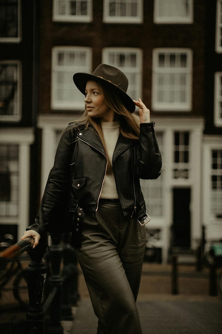 Beautiful female with long hair looking away while standing with hand on hat near metal railing and facade of building on blurred background