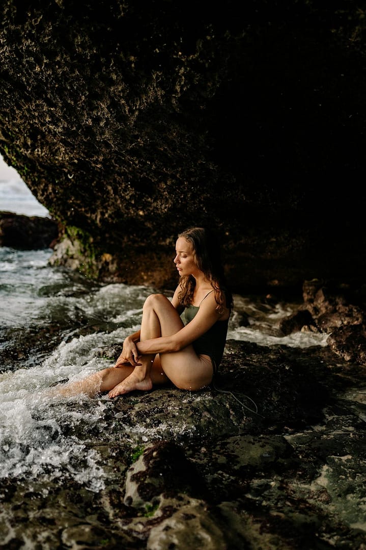 Pensive young woman in swimwear sitting on rocky coast near sea and lowering foot into oncoming wave while looking on sea